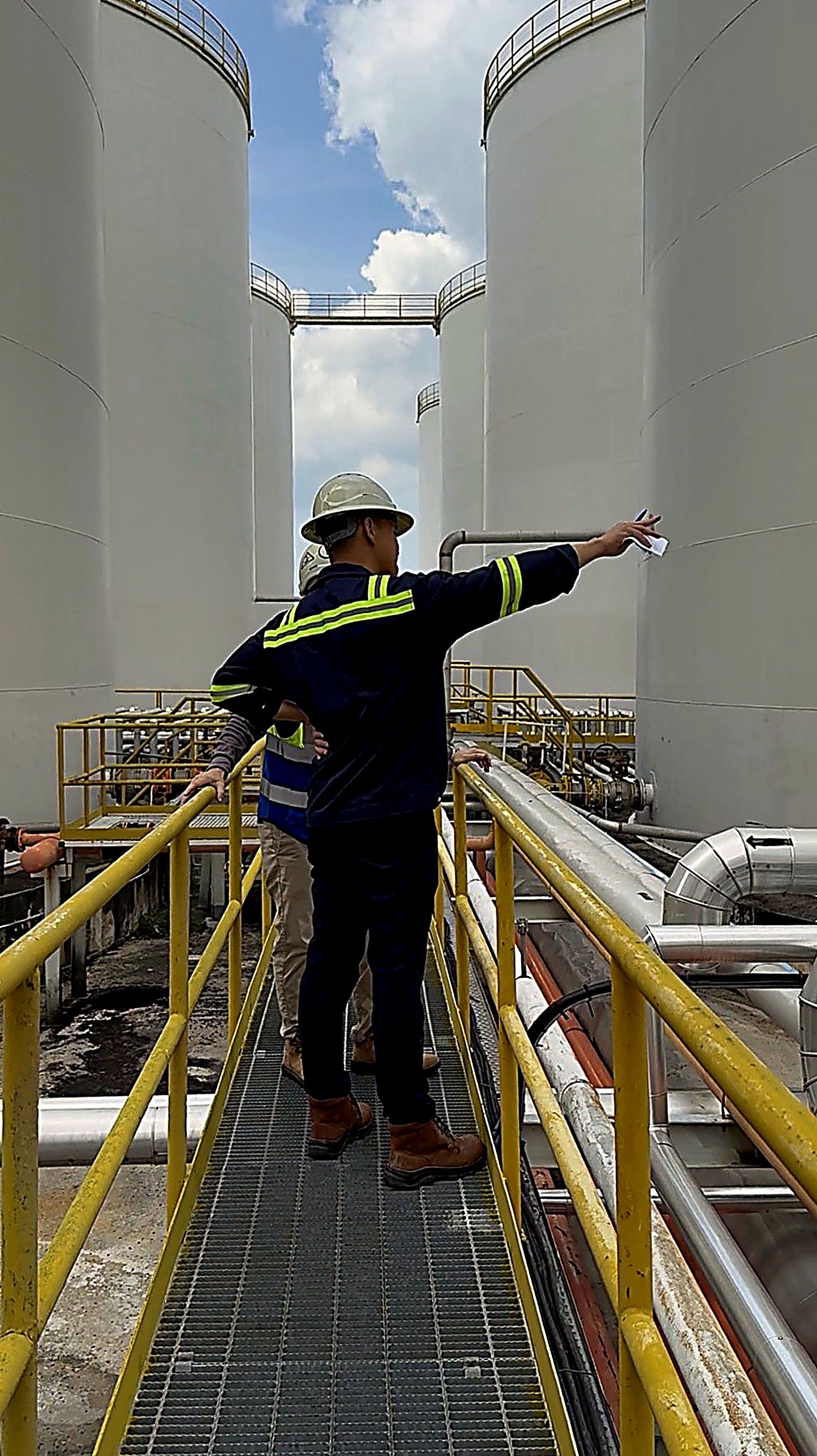 Dean Lai in hard hat and hi-vis on a walkway between storage tanks, pointing out a line to a colleague.